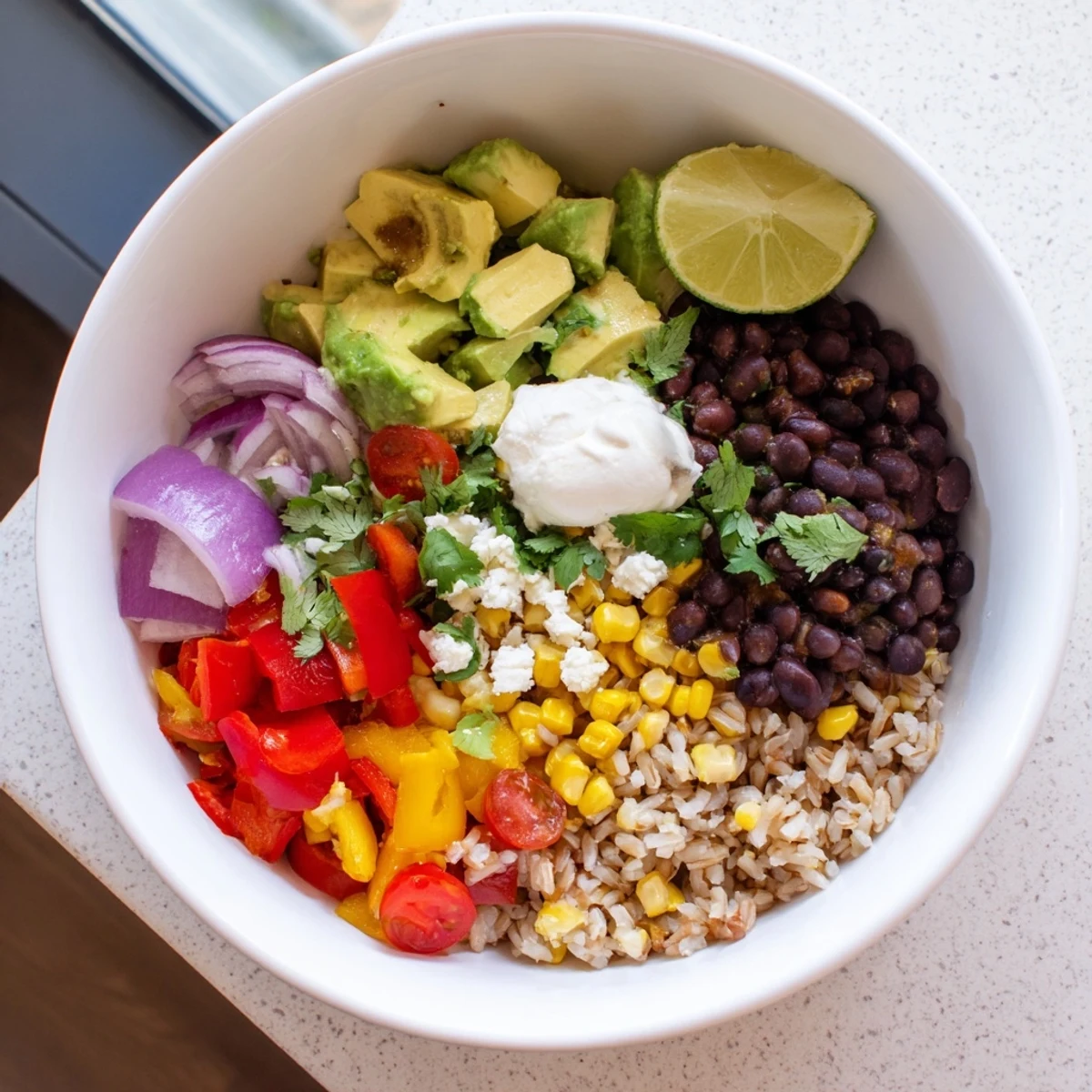 Serving suggestion for a Brown Rice Burrito Bowl with lime wedges, avocado slices, and a dollop of sour cream over colorful ingredients.