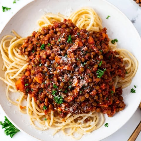 A steaming bowl of lentil bolognese sauce generously spooned over al dente spaghetti, garnished with fresh basil leaves.  